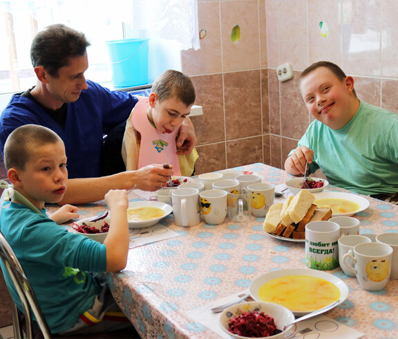 Kinder im Kinderdorf Gomel nehmen gemeinsam mit einem Betreuer ihre Mahlzeiten ein, wobei einige Kinder unterstützt werden.