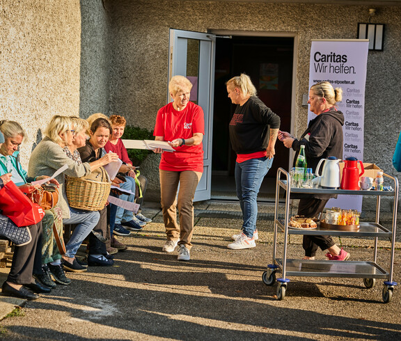 Mehrere Frauen sitzen auf einer Bank und besprechen Dokumente, während eine Caritas-Helferin ihnen Unterlagen überreicht. Im Hintergrund steht ein Tisch mit Getränken und Lebensmitteln sowie ein Caritas-Banner mit der Aufschrift „Caritas Wir helfen“.