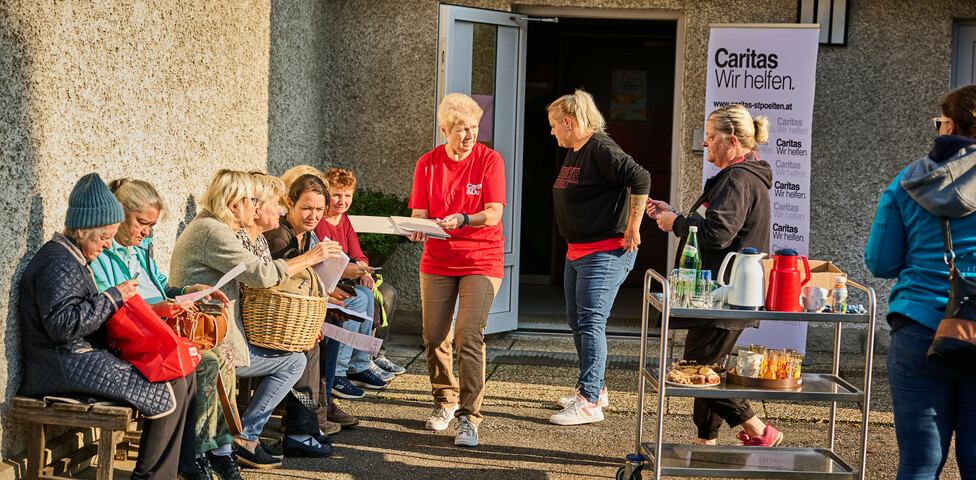 Mehrere Frauen sitzen auf einer Bank und besprechen Dokumente, während eine Caritas-Helferin ihnen Unterlagen überreicht. Im Hintergrund steht ein Tisch mit Getränken und Lebensmitteln sowie ein Caritas-Banner mit der Aufschrift „Caritas Wir helfen“.