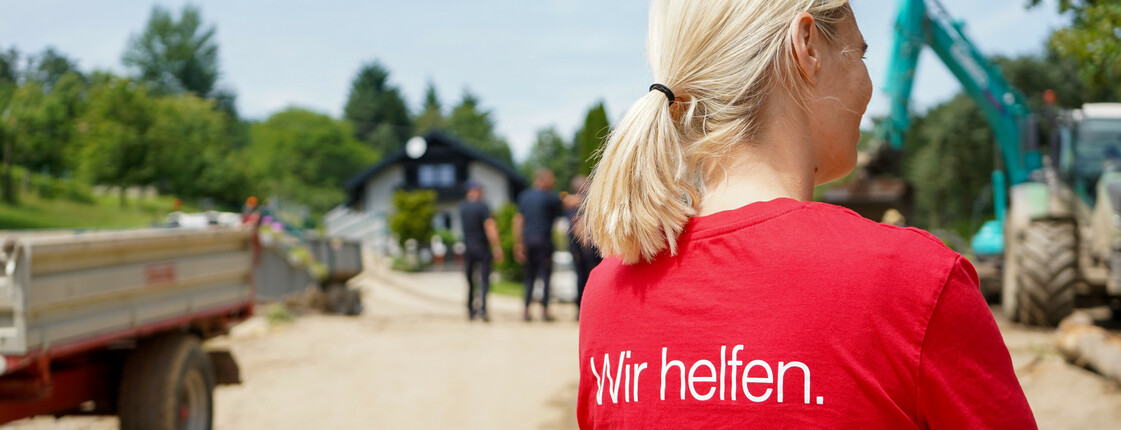 Eine Caritas Mitarbeiterin in einem roten Caritas T-Shirt mit der Aufschrift „Wir helfen“ steht auf einer unbefestigten Straße, während im Hintergrund Bauarbeiten und Menschen vor einem Haus zu sehen sind.