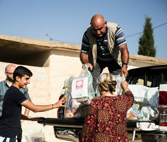 Ein Mann steht auf der Ladefläche eines kleinen LKWs und gibt Säcke mit Gewand an Menschen unten weiter. Auf den Säcken ist das Logo von Caritas Syrien.