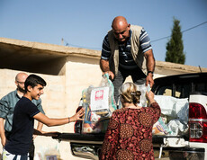 Ein Mann steht auf der Ladefläche eines kleinen LKWs und gibt Säcke mit Gewand an Menschen unten weiter. Auf den Säcken ist das Logo von Caritas Syrien.