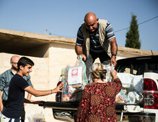 Ein Mann steht auf der Ladefläche eines kleinen LKWs und gibt Säcke mit Gewand an Menschen unten weiter. Auf den Säcken ist das Logo von Caritas Syrien.