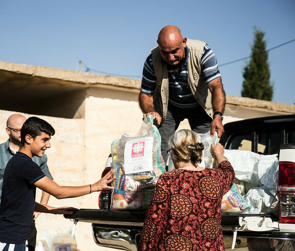 Ein Mann steht auf der Ladefläche eines kleinen LKWs und gibt Säcke mit Gewand an Menschen unten weiter. Auf den Säcken ist das Logo von Caritas Syrien.