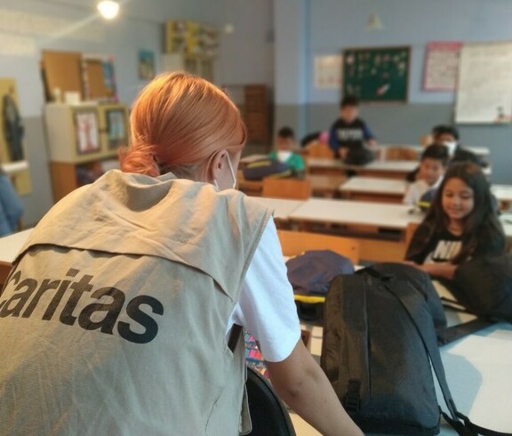 Eine Caritas-Mitarbeiterin ist von hinten zu sehen, sie sieht ins Klassenzimmer, im Hintergrund verschwommen Kinder.