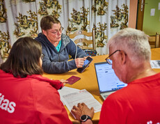 Eine Frau zeigt auf ihrem Smartphone Informationen, während sie mit anderen am Tisch sitzt. Die Gruppe bespricht Hilfeleistungen für Hochwasseropfer in Böheimkirchen. Ein Laptop und ein Reisepass sind auf dem Tisch sichtbar, was auf eine formale Beratung hinweist.