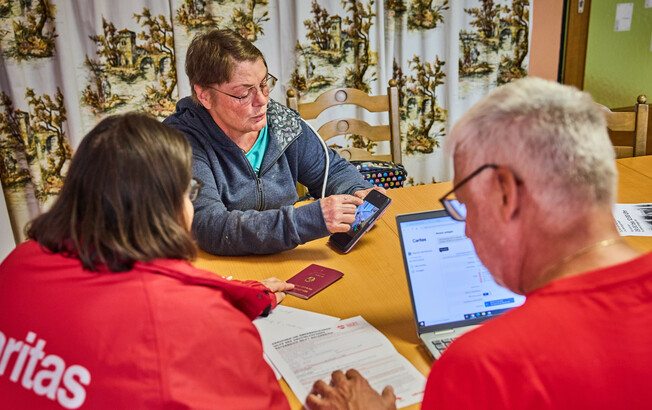 Eine Frau zeigt auf ihrem Smartphone Informationen, während sie mit anderen am Tisch sitzt. Die Gruppe bespricht Hilfeleistungen für Hochwasseropfer in Böheimkirchen. Ein Laptop und ein Reisepass sind auf dem Tisch sichtbar, was auf eine formale Beratung hinweist.