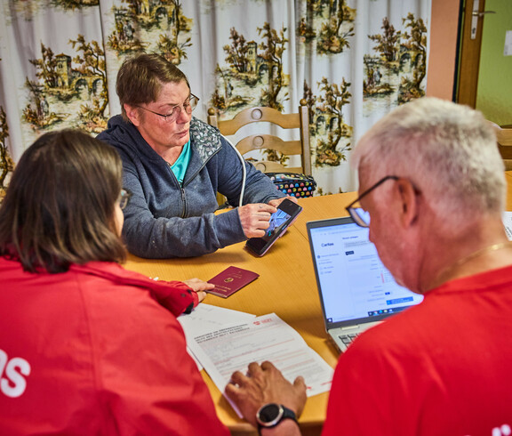 Eine Frau zeigt auf ihrem Smartphone Informationen, während sie mit anderen am Tisch sitzt. Die Gruppe bespricht Hilfeleistungen für Hochwasseropfer in Böheimkirchen. Ein Laptop und ein Reisepass sind auf dem Tisch sichtbar, was auf eine formale Beratung hinweist.