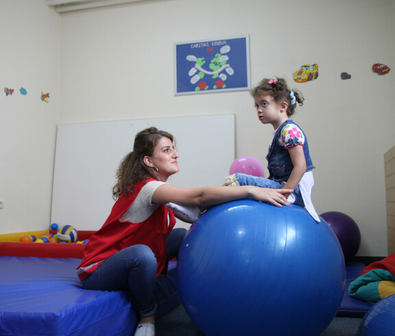 Eine Betreuerin hilft einem Mädchen mit dem Gleichgewicht, während es auf einem Gymnastikball sitzt.