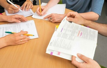 Man sieht die Hände von vier Personen, die an einem Tisch sitzen und Stifte, Zettel und Bücher halten. Die Gesichter sind nicht sichtbar. 