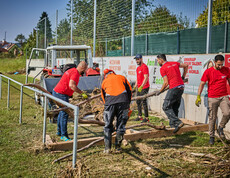 Freiwillige Helfer räumen Baumstämme und Schutt auf einem Sportplatz in Hofstetten-Grünau. Sie tragen rote Caritas-T-Shirts, und im Hintergrund steht ein Traktor bereit, um die Überreste abzutransportieren. Die Helfer arbeiten gemeinsam daran, die Hochwasserschäden zu beseitigen.