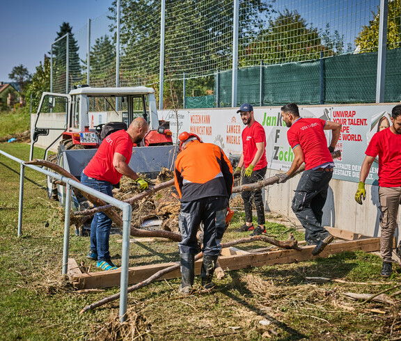 Freiwillige Helfer räumen Baumstämme und Schutt auf einem Sportplatz in Hofstetten-Grünau. Sie tragen rote Caritas-T-Shirts, und im Hintergrund steht ein Traktor bereit, um die Überreste abzutransportieren. Die Helfer arbeiten gemeinsam daran, die Hochwasserschäden zu beseitigen.