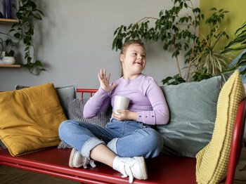 Ein fröhliches Mädchen sitzt entspannt auf einem Sofa, hält eine Tasse und winkt lächelnd.