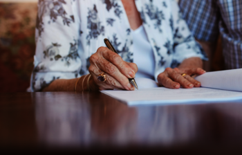 Man sieht die Hände von einer älteren Dame und einem Herren, sie sitzen vor einer Mappe. Die Dame hat einen Stift in der Hand. Die Köpfe der beiden sind nicht erkennbar.