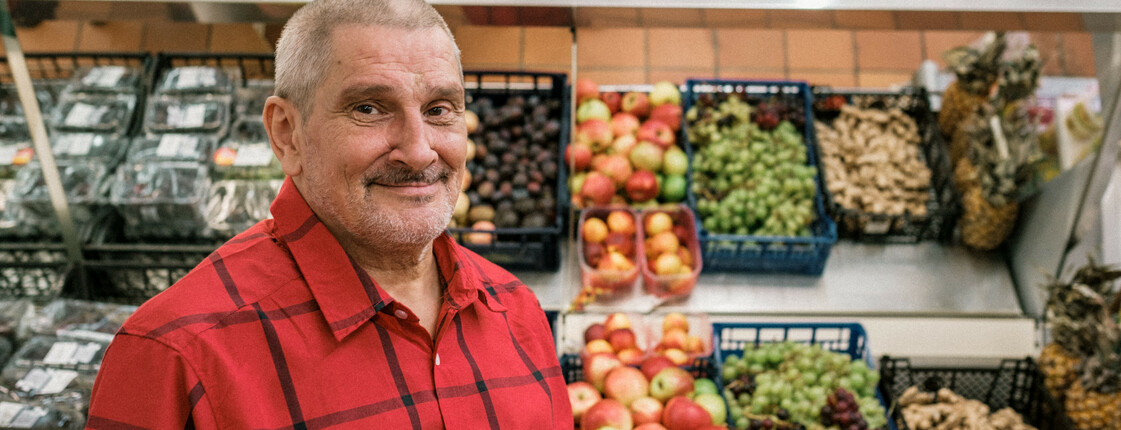 Mann blickt zur Kamera, im Hintergrund ein Obst- und Gemüseregal