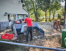 Ein freiwilliger Helfer schaufelt Schlamm in eine Traktorschaufel vor einem Gebäude in Hofstetten-Grünau. Der Mann trägt ein rotes T-Shirt mit der Aufschrift „Wir helfen“. Im Hintergrund sind Bäume und Werkzeuge zu sehen, die für die Aufräumarbeiten verwendet werden.