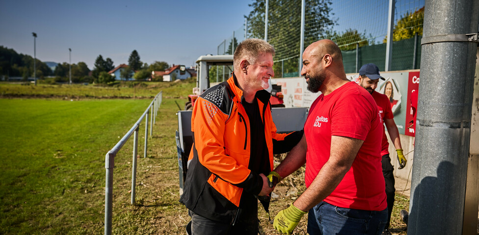 Ein Mann in einer orangen Arbeitsjacke schüttelt lächelnd die Hand eines Freiwilligen in einem roten Caritas-T-Shirt. Im Hintergrund ist ein Traktor und ein Sportplatz zu sehen, wo weitere Helfer an den Aufräumarbeiten beteiligt sind.