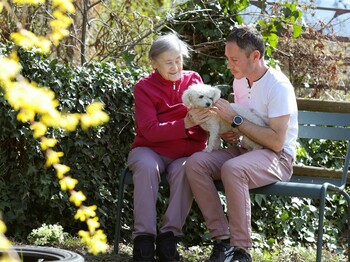 Ältere Dame und Freiwilliger sitzen auf einer Parkbank. Sie haben einen kleinen weißen Hund am Schoß, den sie beide streicheln.