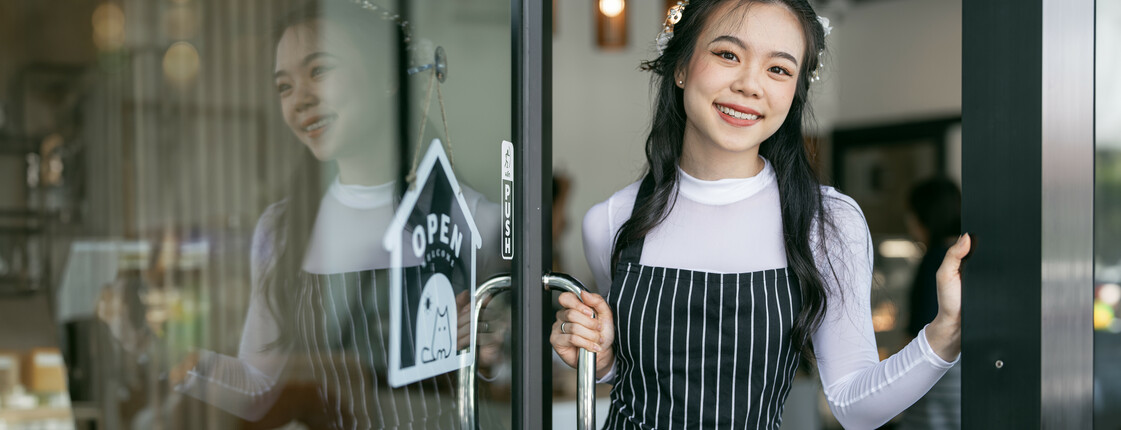 Smiling young waitress waiting for clients at coffee shop. Successful small business owner in blue apron standing at entrance