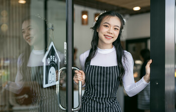 Smiling young waitress waiting for clients at coffee shop. Successful small business owner in blue apron standing at entrance