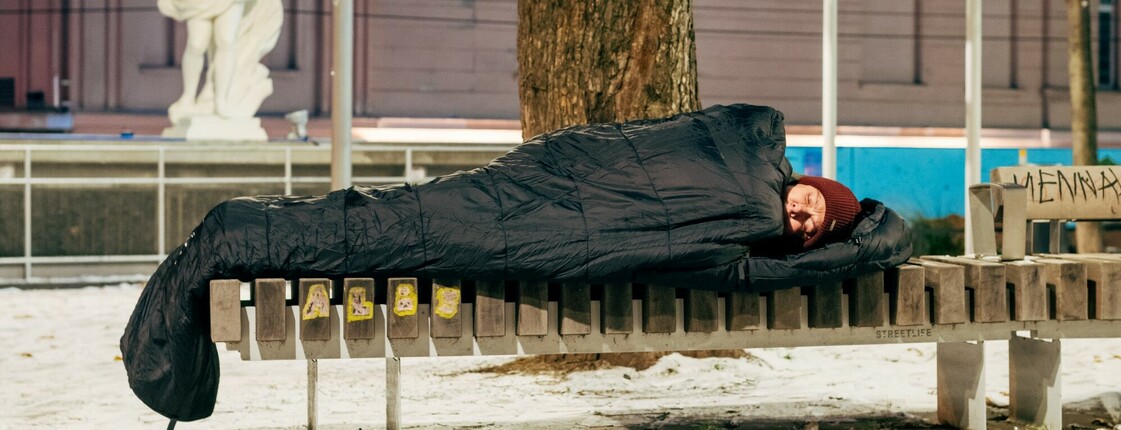 Ein obdachloser Mensch schläft eingehüllt in einen schwarzen Schlafsack auf einer Parkbank im Freien. Der Boden ist mit Schnee bedeckt. 