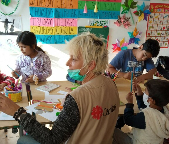 Kinder sitzen rund um einen Tisch und basteln, eine Caritas-Mitarbeiterin sitzt bei ihnen.