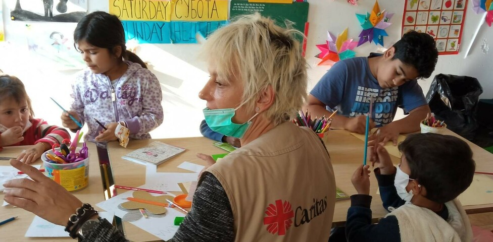 Kinder sitzen rund um einen Tisch und basteln, eine Caritas-Mitarbeiterin sitzt bei ihnen.