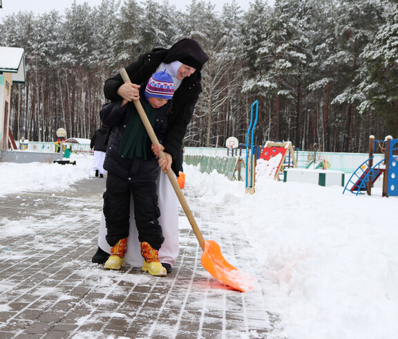 Eine Betreuerin hilft einem Kind beim Schneeschippen auf dem Winterspielplatz im Kinderdorf Gomel, umgeben von Spielgeräten und Schnee.