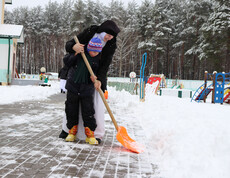 Eine Betreuerin hilft einem Kind beim Schneeschippen auf dem Winterspielplatz im Kinderdorf Gomel, umgeben von Spielgeräten und Schnee.