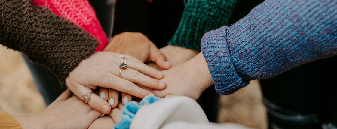 Mehrere Hände unterschiedlicher Personen liegen übereinander als Zeichen von Gemeinschaft und Zusammenhalt. Die Personen tragen warme, bunte Strickpullover.