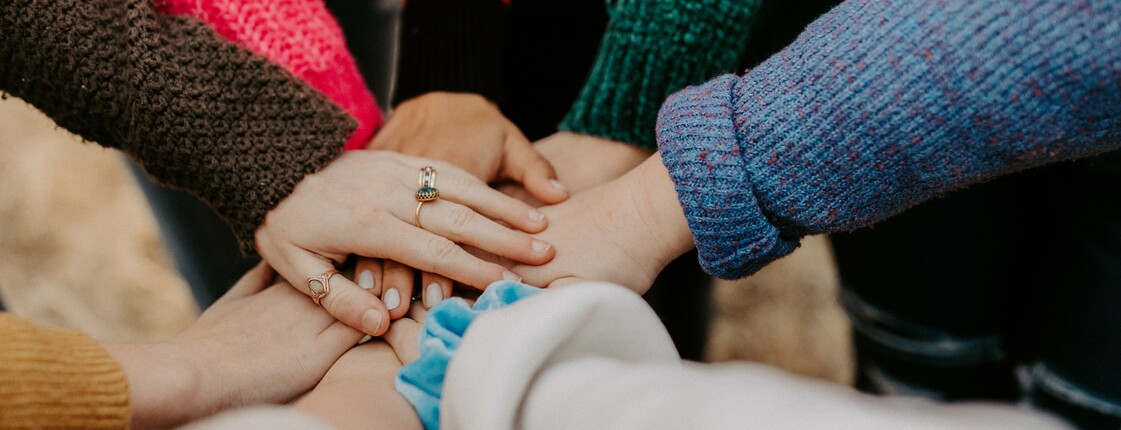 Mehrere Hände unterschiedlicher Personen liegen übereinander als Zeichen von Gemeinschaft und Zusammenhalt. Die Personen tragen warme, bunte Strickpullover.