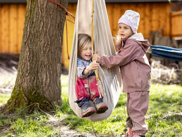 Zwei Kindergartenkinder in Regenkleidung spielen im Garten mit einer Stoff-Schaukel.