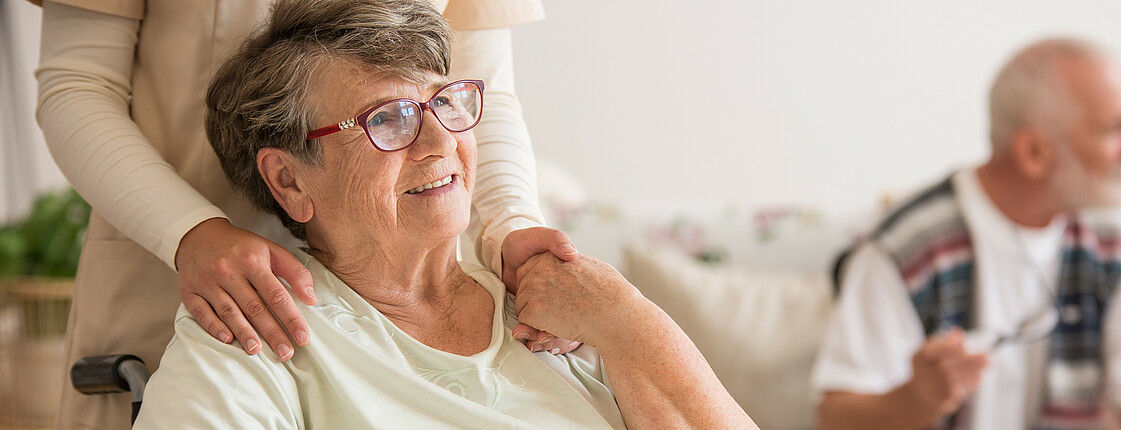 Eine Seniorin mit Brille sitzt in einem Rollstuhl und hält die Hand einer Angehörigen, welche hinter ihr steht.