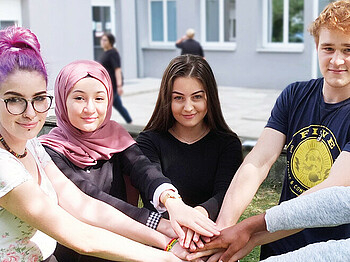 3 Schülerinnen und 2 Schüler stehen im Halbkreis beieinander im Grünen im Hof der Schule und Halten die Hände übereinander. Im Hintergrund steht verschwommen eine Schülerin mit Buch in der Hand. Copyright Caritas Schule Wiener Neustadt