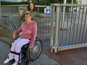 Zwei Frauen vor einem halb geöffneten Eisentor, eine der beiden sitzt im Rollstuhl. Beide lächeln.