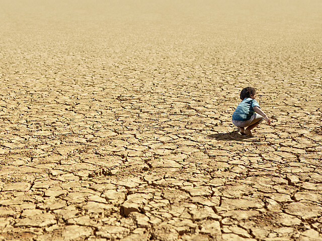 ein kleines Kind hockt barfuß in einer ausgetrockneten Landschaft, welche bis zum Horizont von Dürre gezeichnet ist