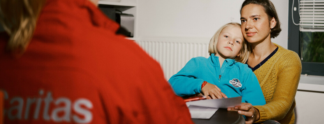 Eine Mutter sitzt mit ihrem Kind auf dem Schoß. Ihnen gegenüber am Tisch sitzt eine Frau mit einer roten Caritas-Jacke, sie ist von hinten zu sehen. 