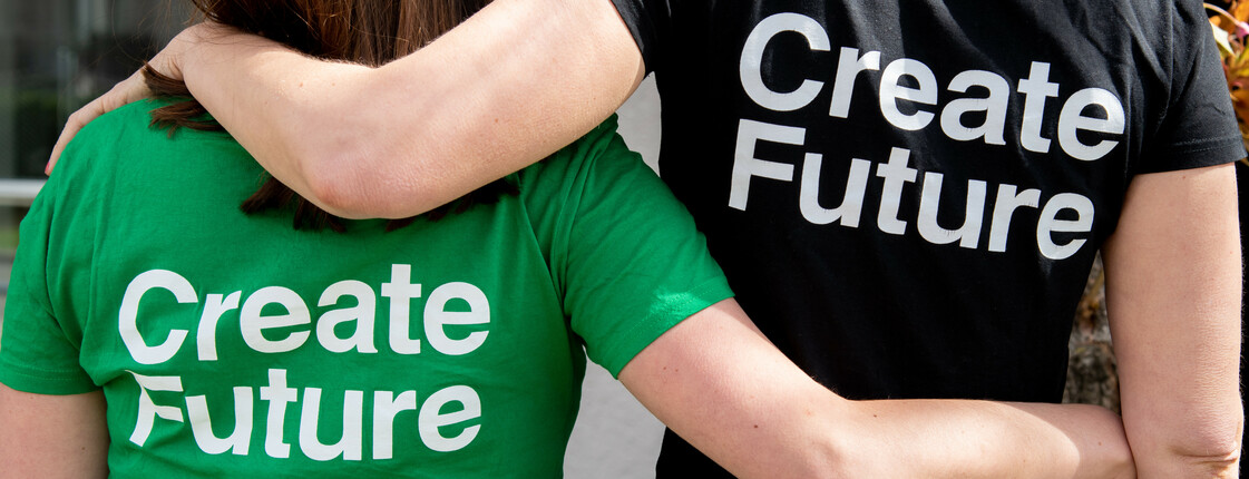 Man sieht zwei Personen von hinten, sie haben die Arme auf ihren Schultern und tragen T-Shirts mit der Aufschrift "Create Future".