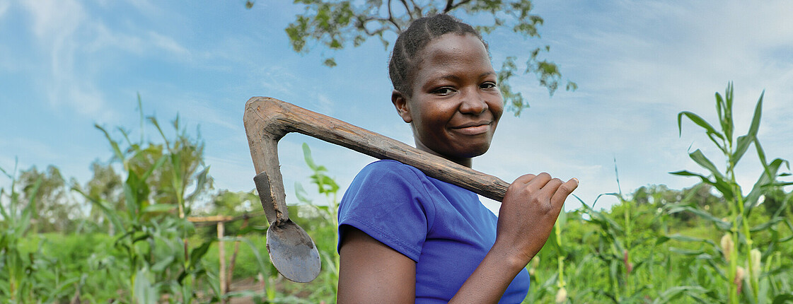 Eine junge Frau steht auf einem grünen Feld und trägt eine Hacke über ihrer Schulter. Sie lächelt in die Kamera. Das Bild steht im Kontext der Hungerkampagne 2024.