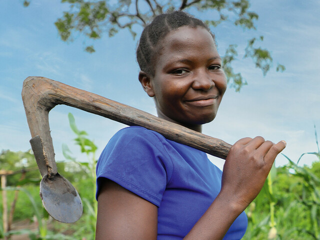 Eine junge Frau steht auf einem grünen Feld und trägt eine Hacke über ihrer Schulter. Sie lächelt in die Kamera. Das Bild steht im Kontext der Hungerkampagne 2024.